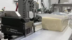 Matthews Marking Systems En L-serie- Bläckstråle -skrivare placerad bredvid en plastförpackad produkt på ett transportband.