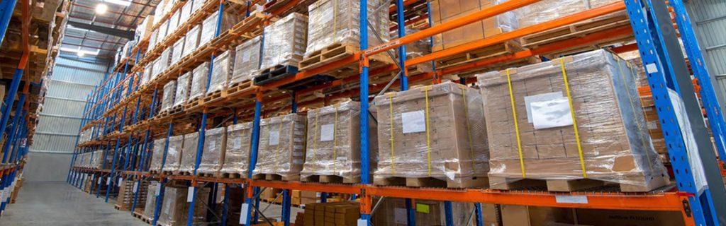 Rader av krympplastförpackade pallar med kartonger, uppställda på industriella lagerhyllor för transport och distribution.